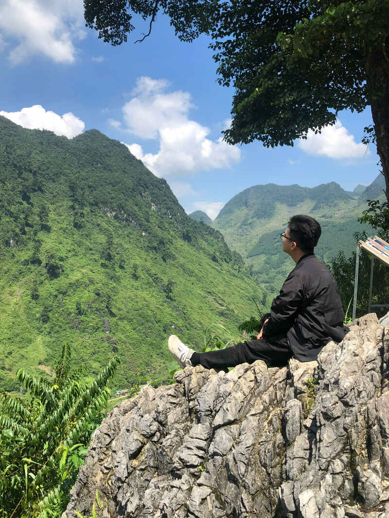 I’m sitting on a rock under the Lonely Tree viewpoint on the Ha Giang Loop, Vietnam, admiring the green mountain range.